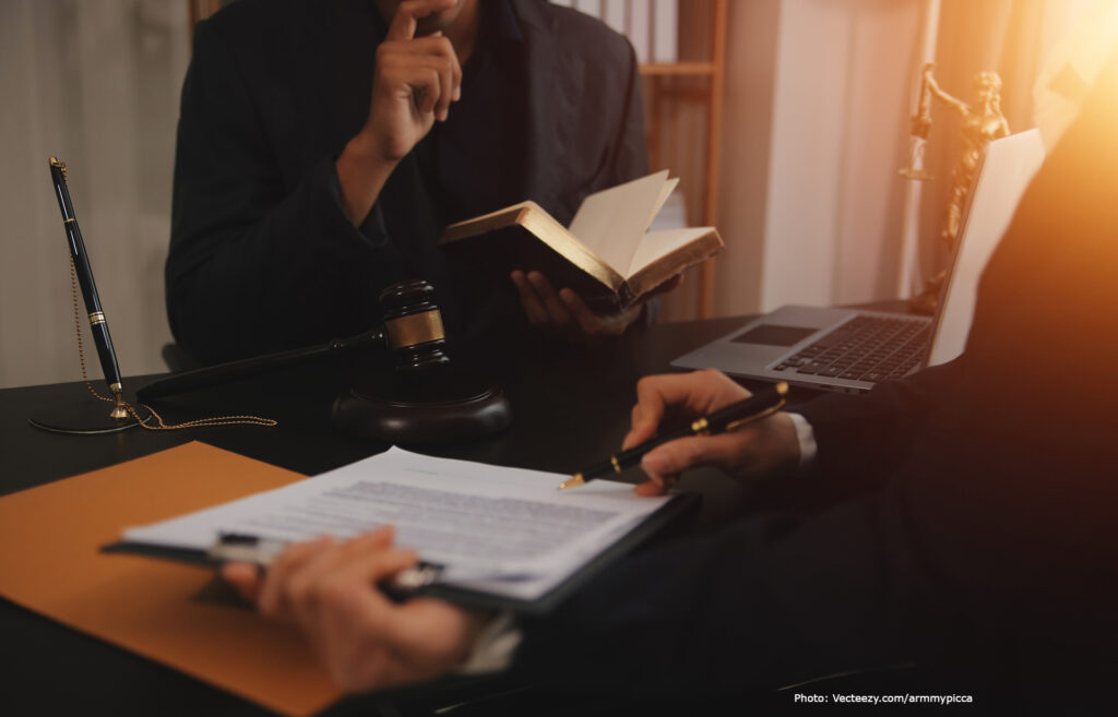Osoba trzymająca kodeks prawny, zastanawiająca się, obok młotek, laptop oraz druga osoba trzymająca dokumenty i pióro – symbolika pracy prawniczej i studiowania prawa
