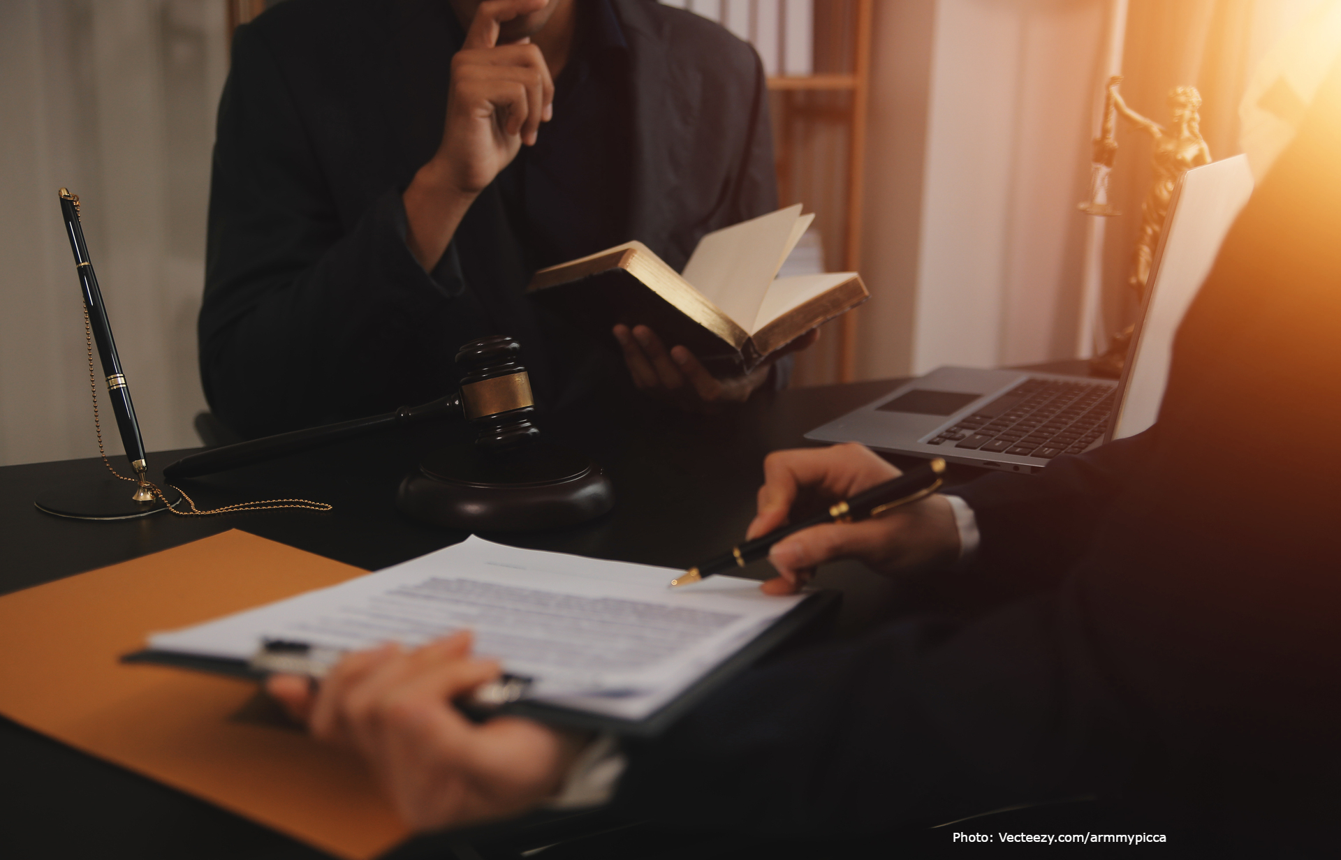 Osoba trzymająca kodeks prawny, zastanawiająca się, obok młotek, laptop oraz druga osoba trzymająca dokumenty i pióro – symbolika pracy prawniczej i studiowania prawa